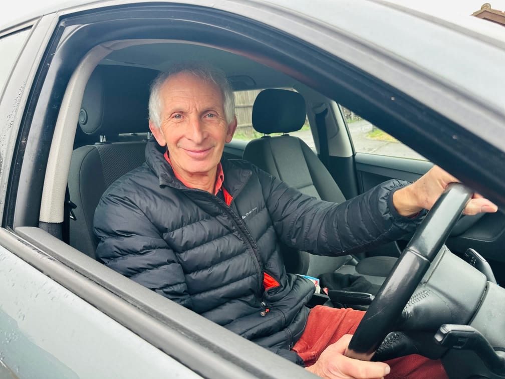 A man sits in the car with his hands on the steering wheel, he looks at the camera and smiles, he is wearing a jacket and red collar. The rain in the background makes rain drops on the window and the car is grey in colour.